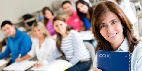 A woman stands in front of a classroom holding a notebook with the letters CCHC.